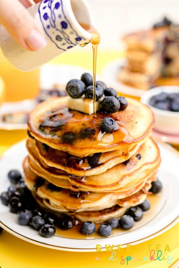 Hand pouring syrup over stacked Fluffy Blueberry Pancakes on white plate.