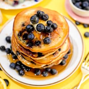 Square image of pancakes stacked topped with blueberries and syrup.
