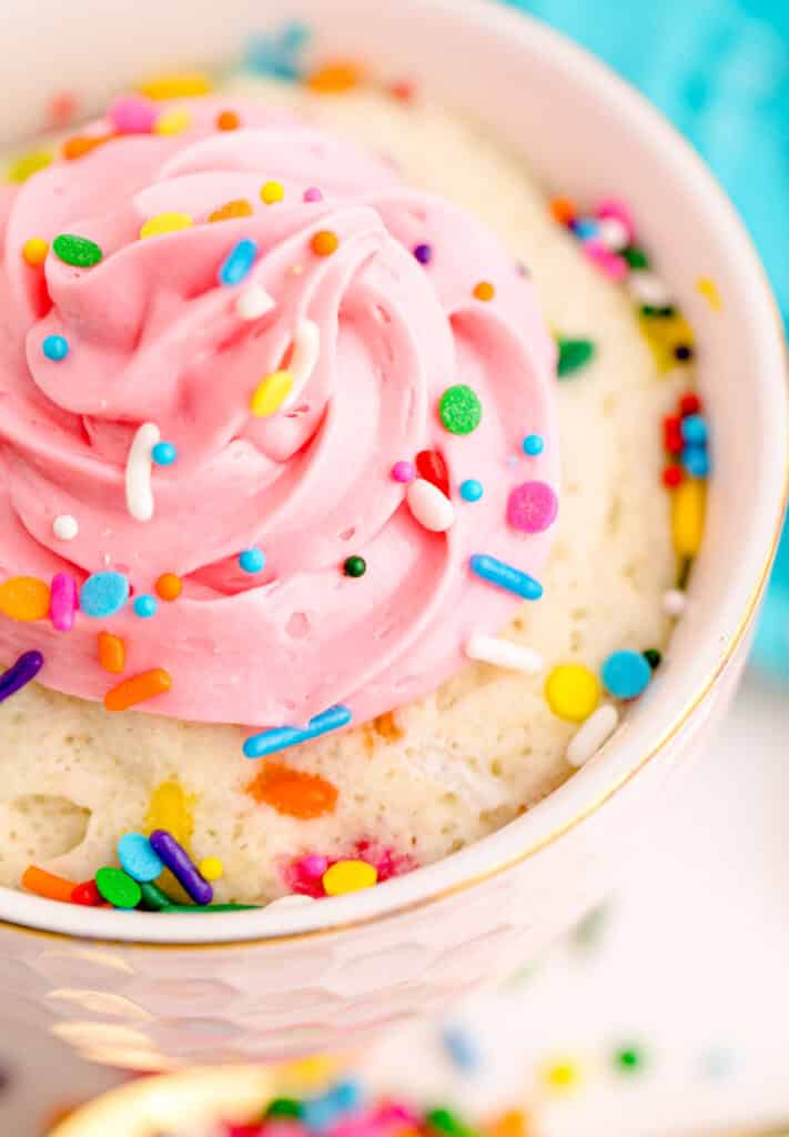 A close up image of a mug cake with cake mix, sprinkles and pink frosting