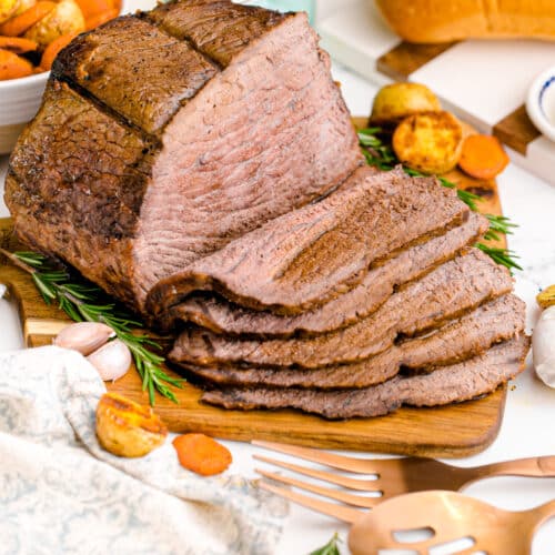 Rump roast partially carved sitting on a sutting board with vegetables and bread on the table.