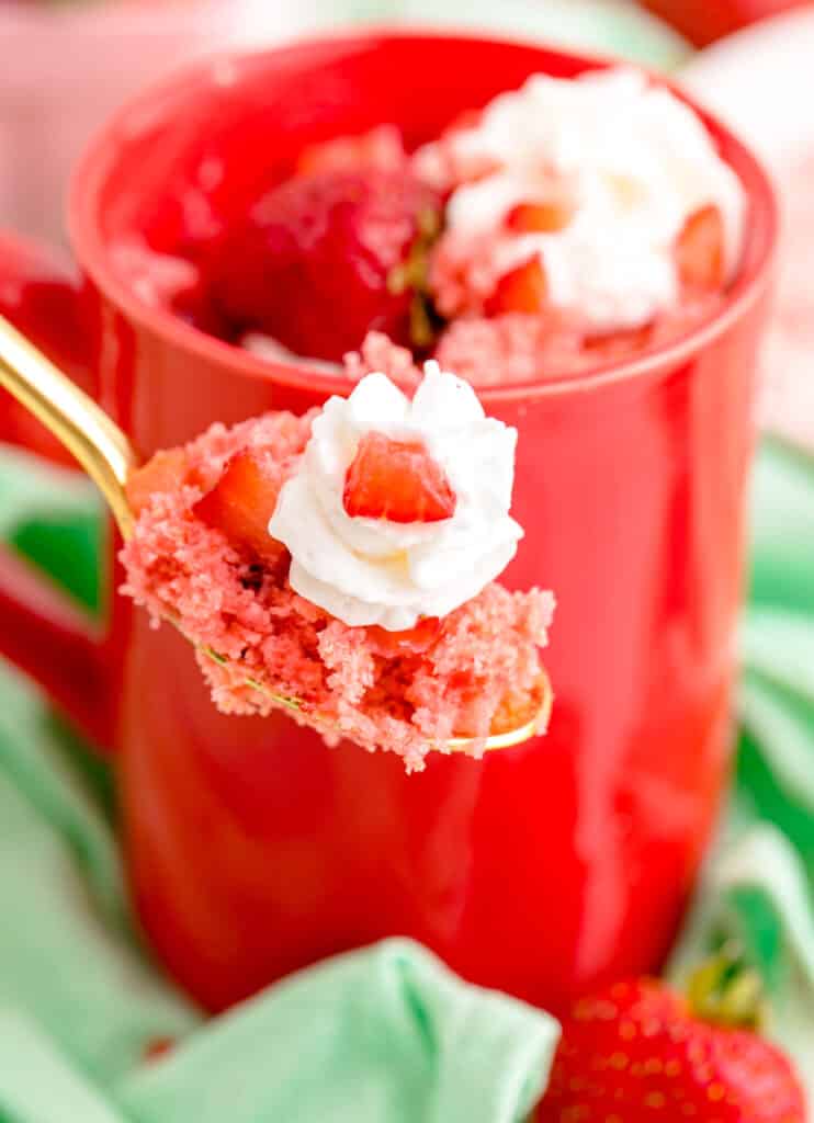 A spoonful of strawberry mug cake with whipped cream and diced strawberries on a gold spoon.