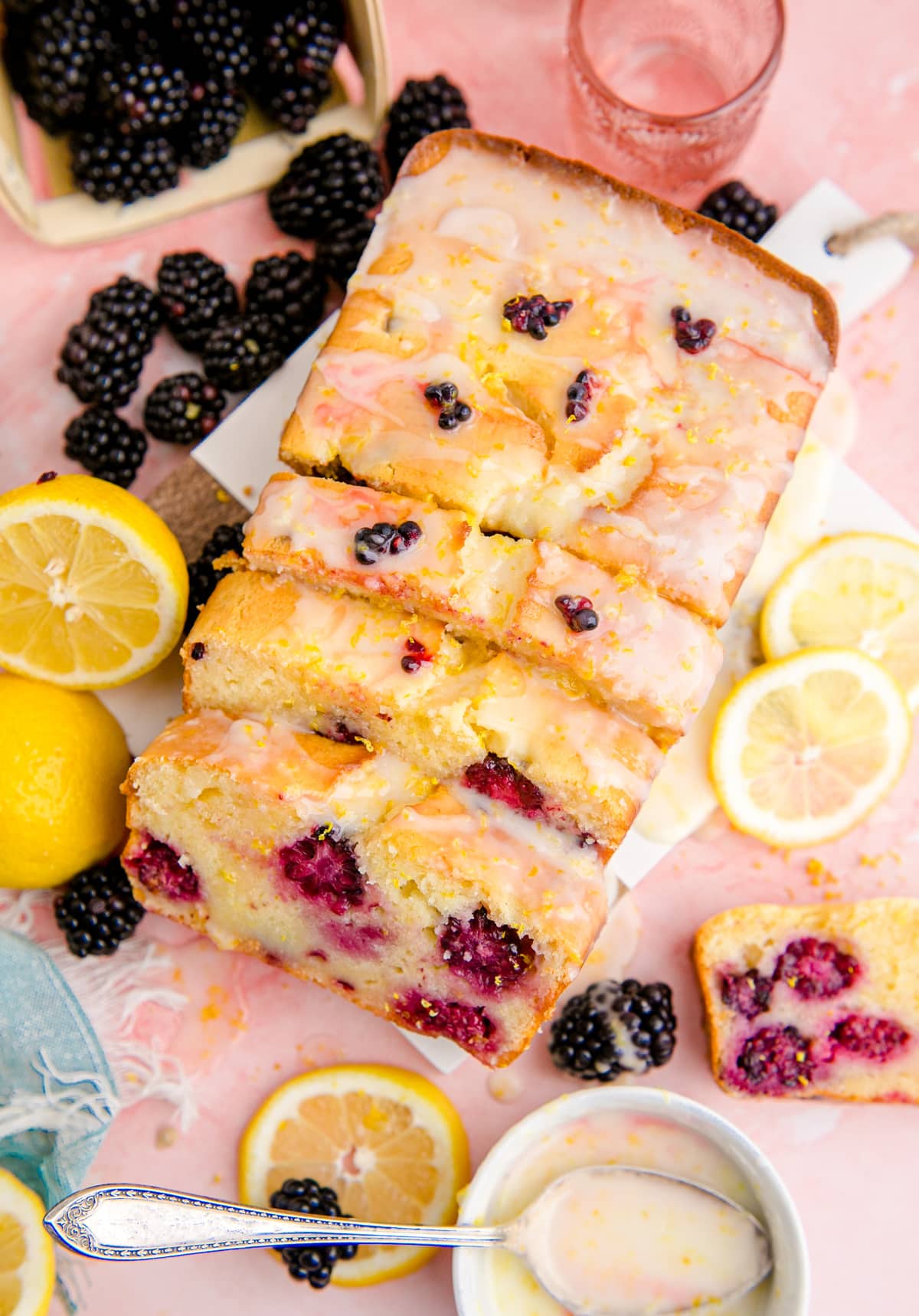 Blackberry Lemon Bread loaf cut into slices and glazed on a white cutting board and surrounded by fresh blackberries and lemons.