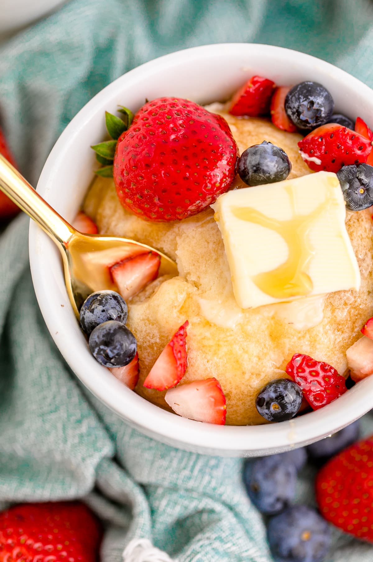 Close up photo of a gold spoon scooping out a helping of pancakes with berries and syrup out of a white mug.