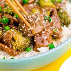 Close up photo of a thin slice of cooked meat a being picked up with chopsticks from a bowl of beef and broccoli over rice.