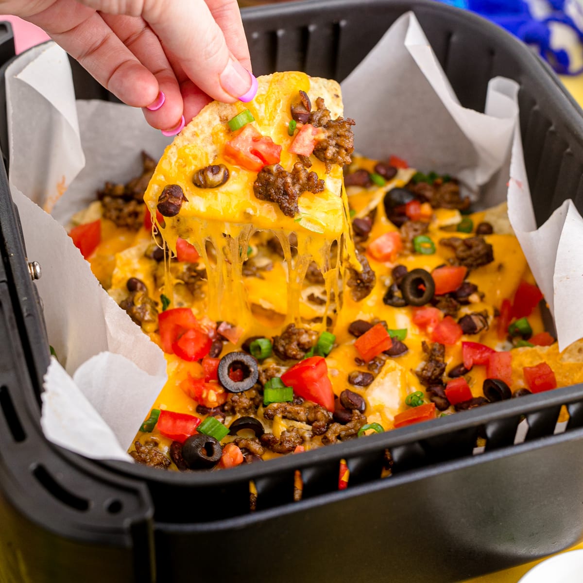 A tortilla chip being with melty cheese and toppings being lifted out an air fryer full of nachos.