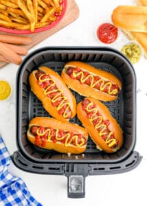 Hot dogs in an air fryer basket in buns and topped with ketchup and mustard.