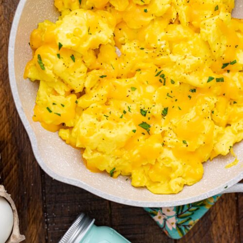 Fluffy scrambled eggs topped with cheese, ground black pepper, parsley in a pan on a wooden countertop.