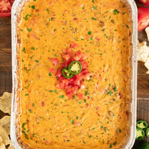 Smoked queso in a 9 x 13 disposable foil pan topped with parsley, diced tomatoes and sliced jalapeños.