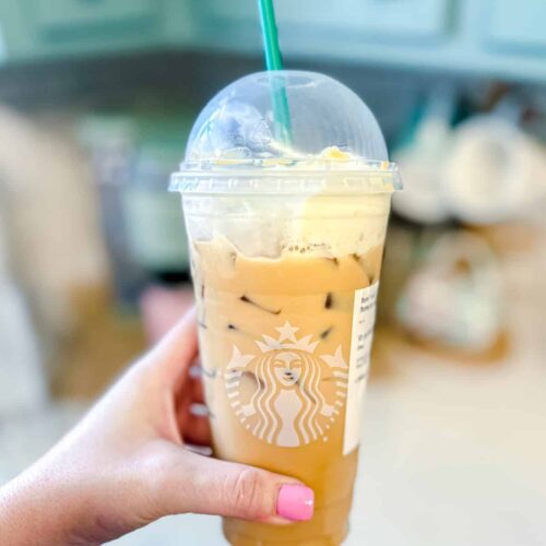 A hand holding a Venti Starbucks cup with an iced white mocha and it topped with whipped cream.
